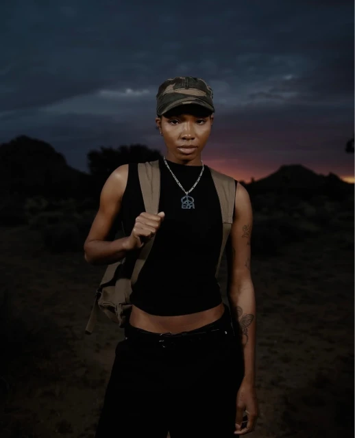 Woman in a black tank top and camo hat poses outdoors against a dusky sky with mountains. She wears a tan vest and necklace.