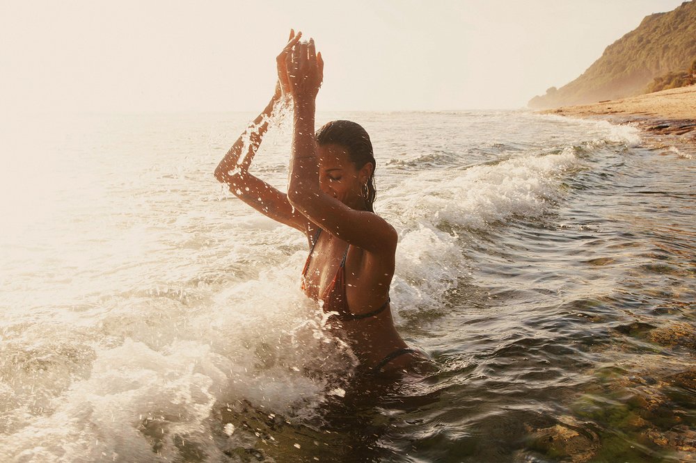 Woman in a bikini standing in ocean surf with hands raised above her head, splashing water against a sunlit sandy beach.