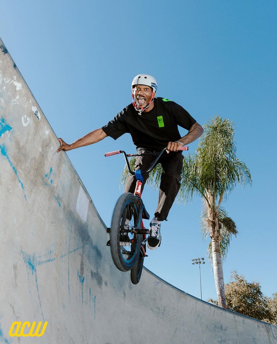A Black male BMX rider is mid-air on a bike inside a concrete skatepark, wearing a helmet, black t-shirt, and pants, under a clear blue sky.