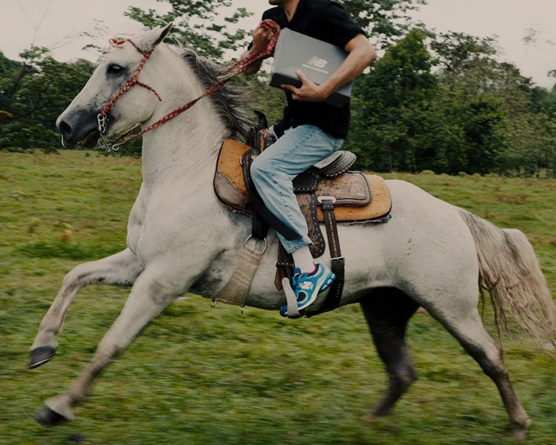 Man rides a galloping white horse across a grassy field carrying a New Balance shoebox in an outdoor daytime setting.