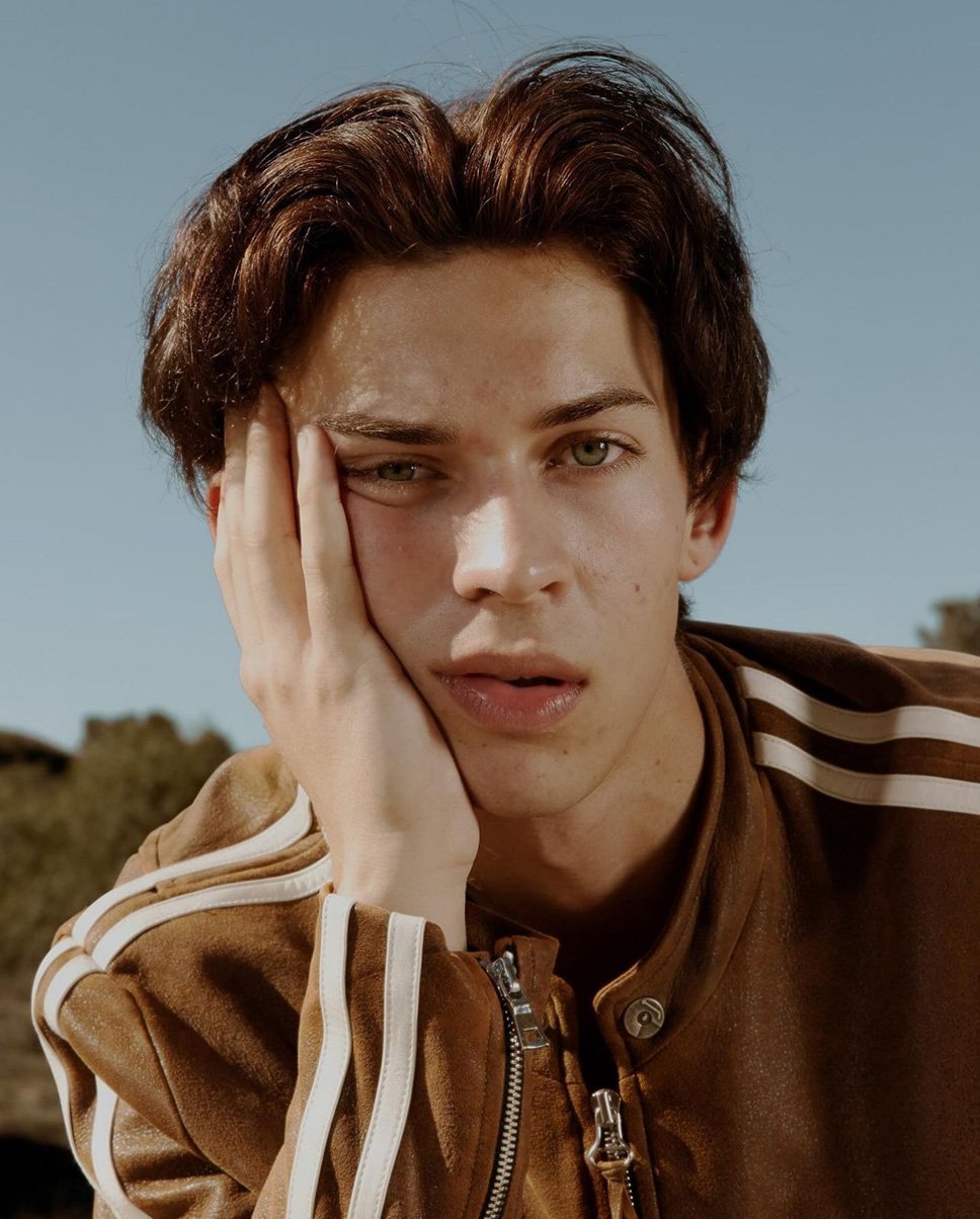 Close-up portrait of a young adult male with brown hair, wearing a brown jacket with white stripes, posing outdoors against a clear blue sky.