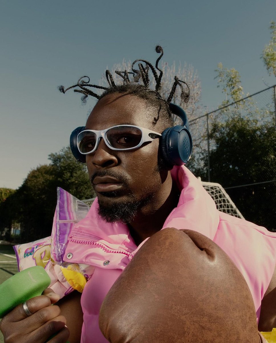 Adult male outdoors wears sunglasses and headphones, with boxing gloves and snacks, on a tennis court under a blue sky.