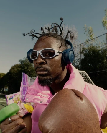 Adult male outdoors wears sunglasses and headphones, with boxing gloves and snacks, on a tennis court under a blue sky.
