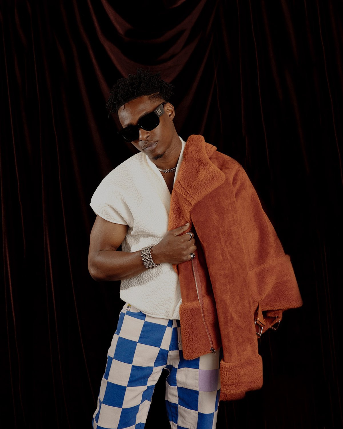 Portrait of a young Black man in patterned pants and sunglasses with an orange jacket draped over his shoulder in front of curtains.
