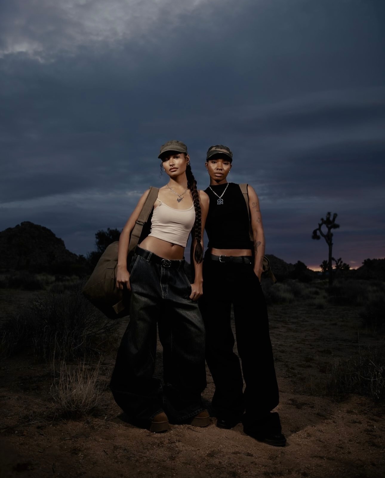 Two models standing outdoors in a desert landscape under a stormy dusk sky, wearing caps and casual streetwear.
