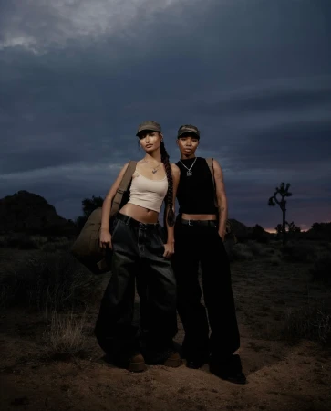 Two models standing outdoors in a desert landscape under a stormy dusk sky, wearing caps and casual streetwear.
