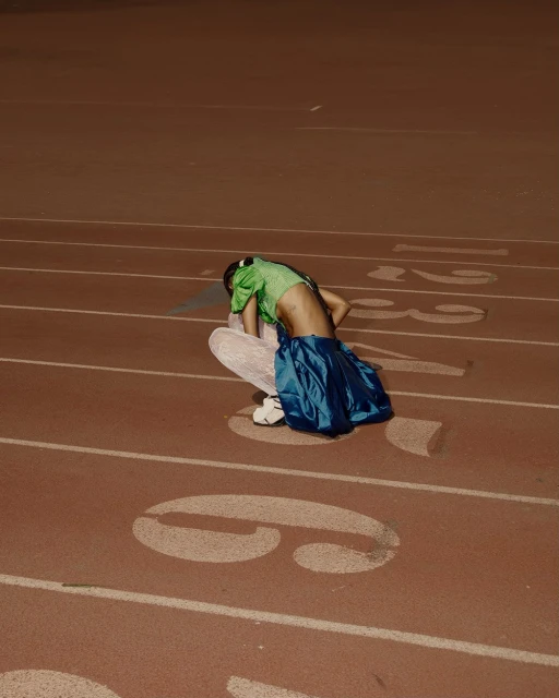 Female athlete in green top and blue skirt crouches on red running track with white lines and painted numbers in outdoor setting.