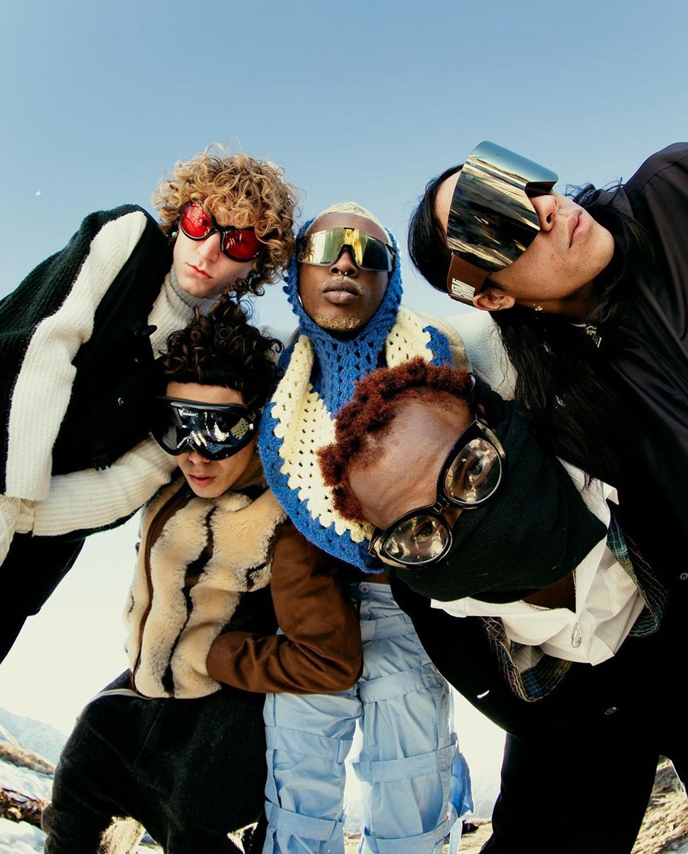 Five people wearing futuristic ski goggles pose closely together outdoors on a sunny day, with a bright blue sky and snow visible.