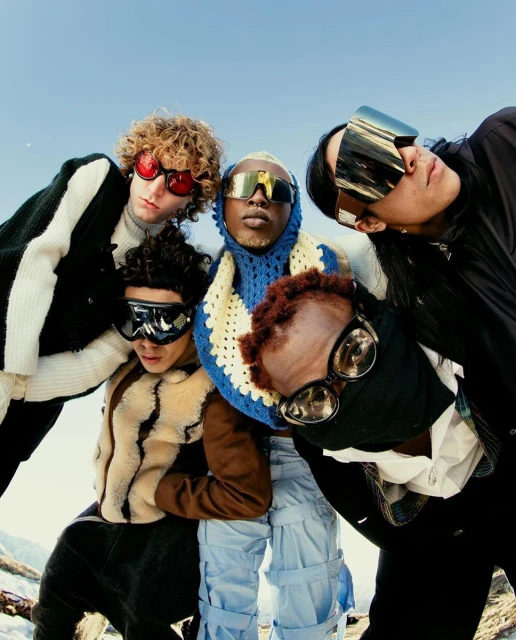 Five people wearing futuristic ski goggles pose closely together outdoors on a sunny day, with a bright blue sky and snow visible.