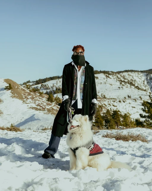 Person with red hair and sunglasses, wearing a black coat, posing with a Samoyed dog wearing a red sweater, in a snowy mountain landscape.