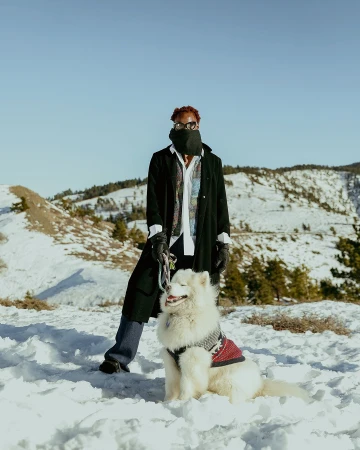 Person with red hair and sunglasses, wearing a black coat, posing with a Samoyed dog wearing a red sweater, in a snowy mountain landscape.