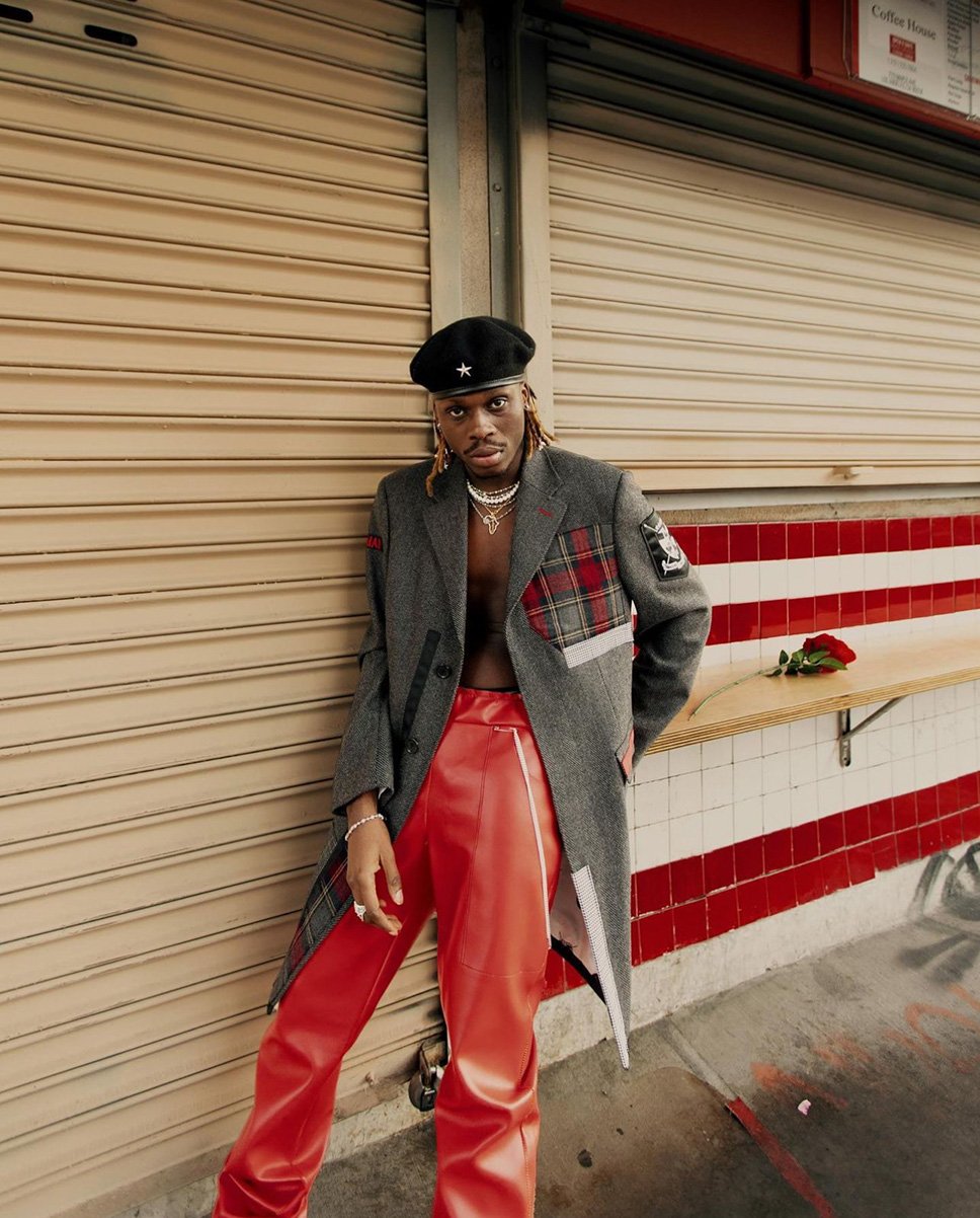 A young adult with dreadlocks and beret posing against closed shutters in a grey coat and red pants with a red rose nearby.