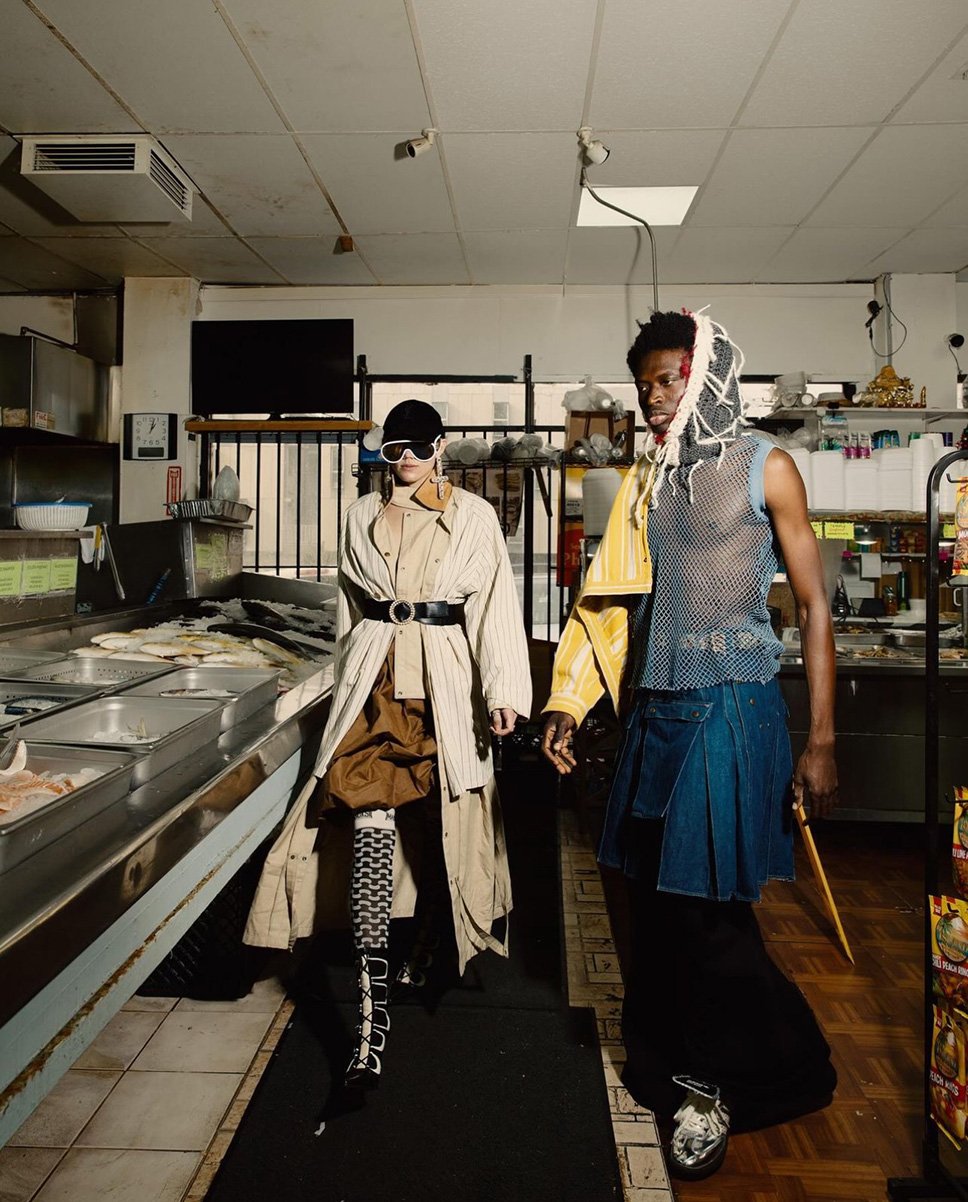 Two fashionably dressed people in an urban market interior; one in a trench coat and the other in avant-garde streetwear fashion.