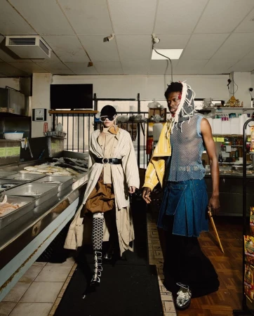 Two fashionably dressed people in an urban market interior; one in a trench coat and the other in avant-garde streetwear fashion.