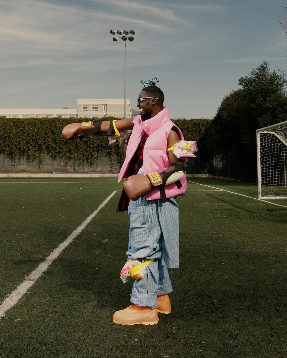 Man in a pink vest and blue striped pants stands on a green field with soccer goals, holding boxing gloves and gesturing.