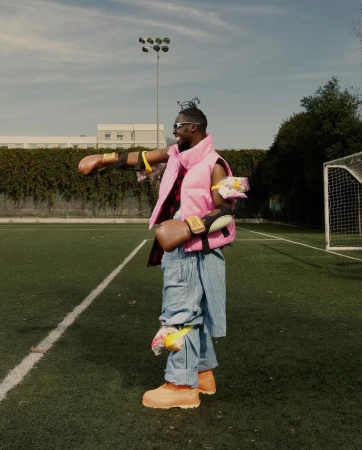 Man in a pink vest and blue striped pants stands on a green field with soccer goals, holding boxing gloves and gesturing.