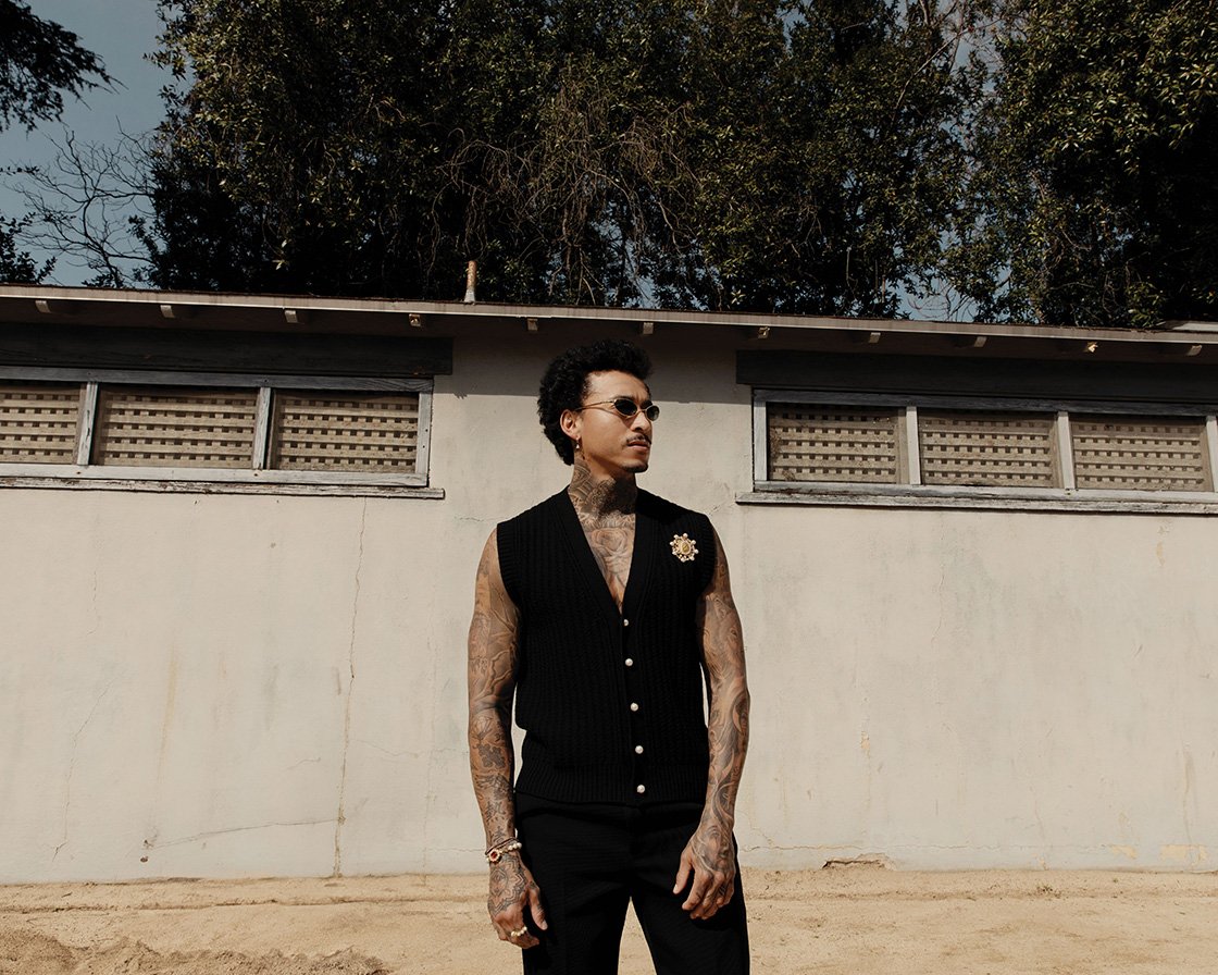 Adult male poses in a black vest and pants outdoors in front of a white building with trees in the background on a sunny day.