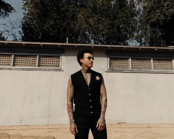 Adult male poses in a black vest and pants outdoors in front of a white building with trees in the background on a sunny day.