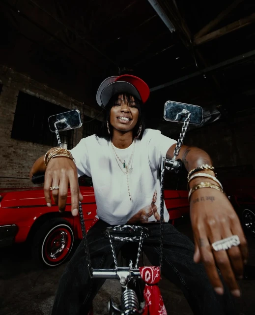 A woman with a backwards baseball cap poses on a lowrider bicycle in front of a red car inside a dimly lit auto body shop.