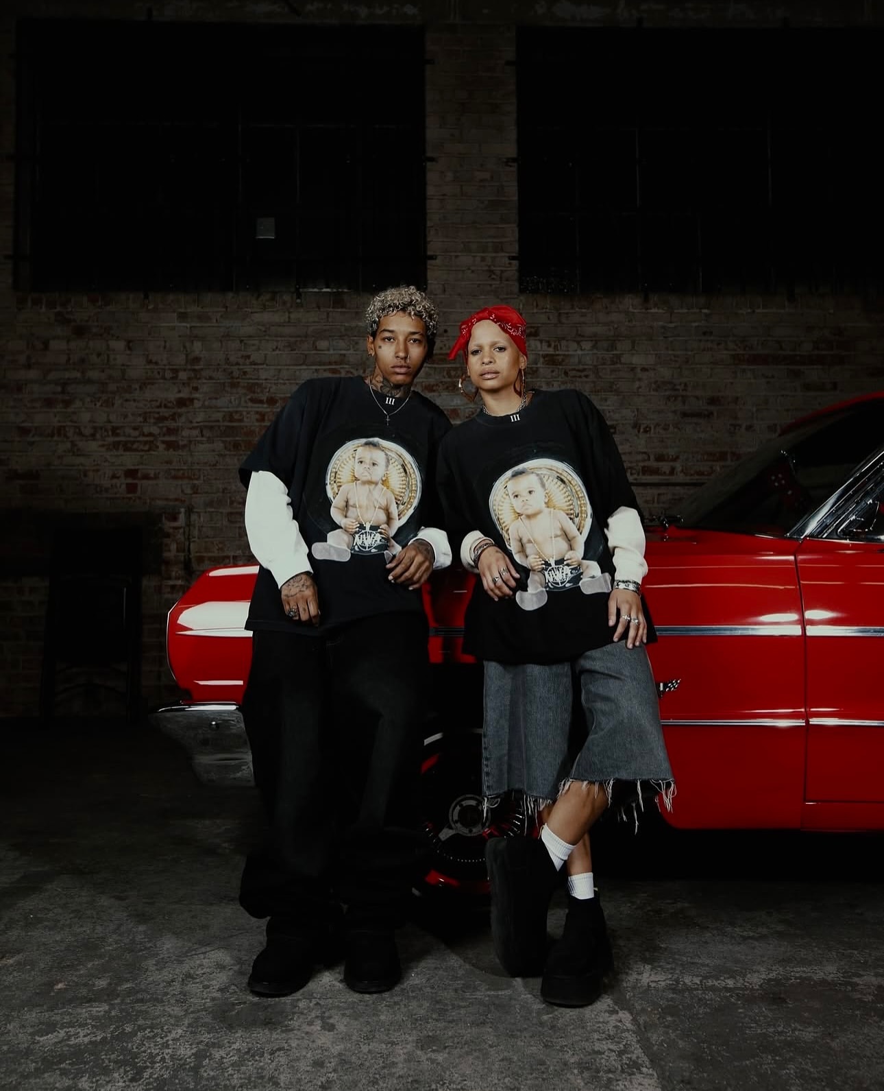 Two adult models pose with a red vintage car inside an industrial-style building, wearing matching shirts with graphic prints.