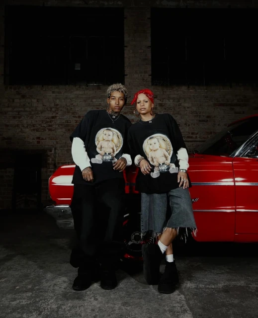 Two adult models pose with a red vintage car inside an industrial-style building, wearing matching shirts with graphic prints.