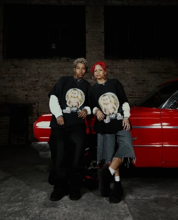 Two adult models pose with a red vintage car inside an industrial-style building, wearing matching shirts with graphic prints.