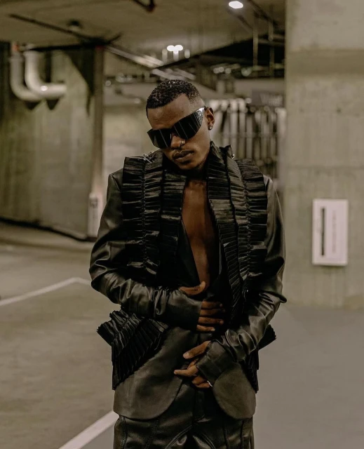 An adult male poses in a futuristic black jacket and pants, standing in a dimly lit parking garage with concrete walls and pipes.