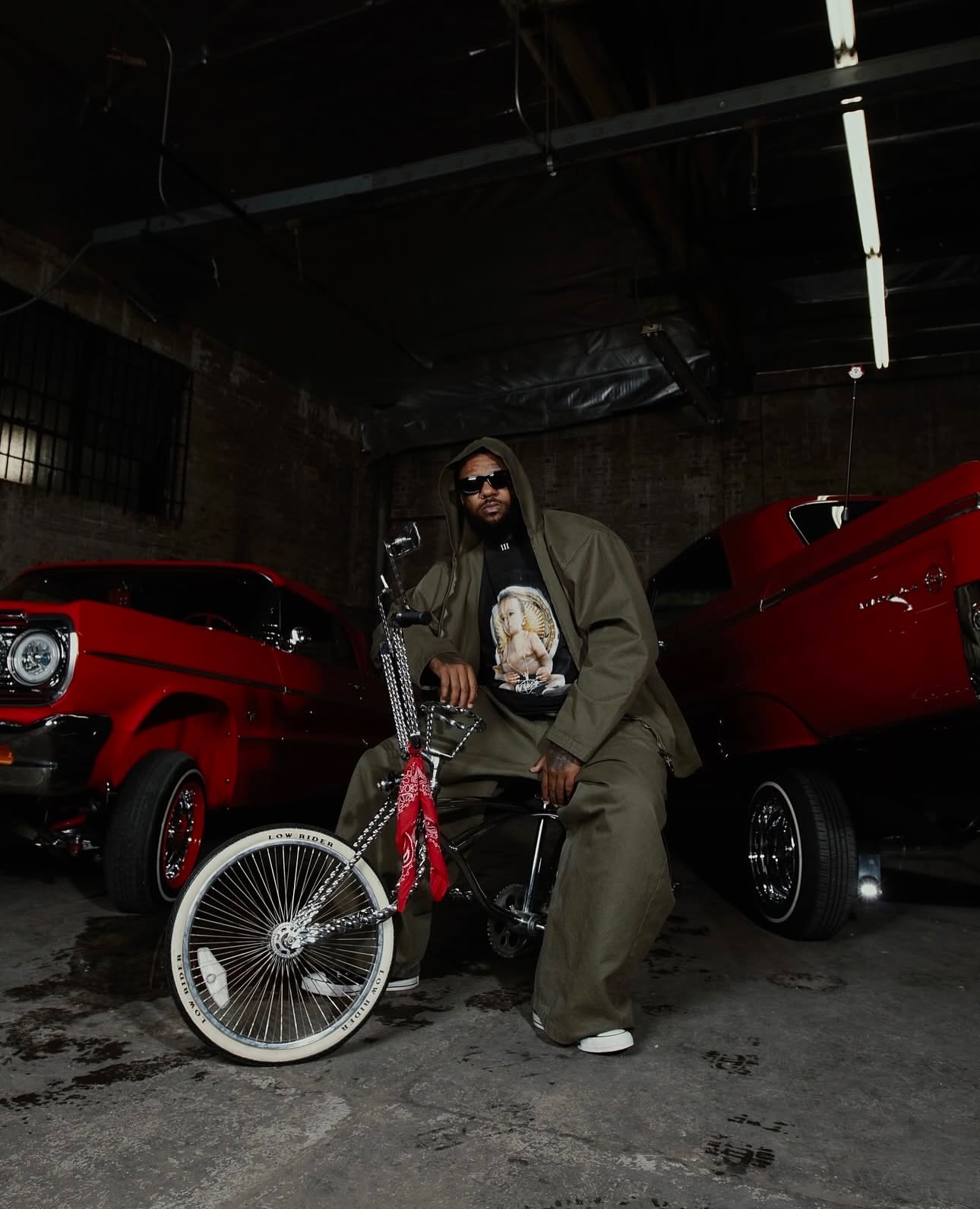 Man in an olive green suit poses with a lowrider bike, flanked by two red vintage cars in a dimly lit, gritty, indoor setting.