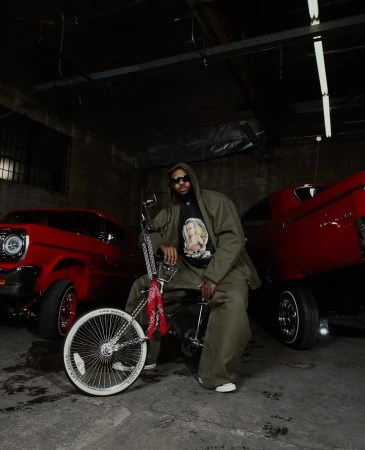 Man in an olive green suit poses with a lowrider bike, flanked by two red vintage cars in a dimly lit, gritty, indoor setting.