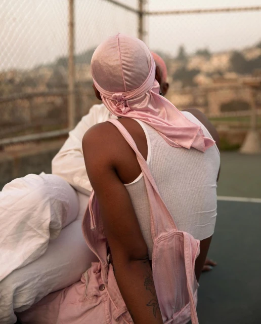 Two people sit outdoors, wearing pink and white clothing, one with a pink head covering, at eye-level on a sunny day.