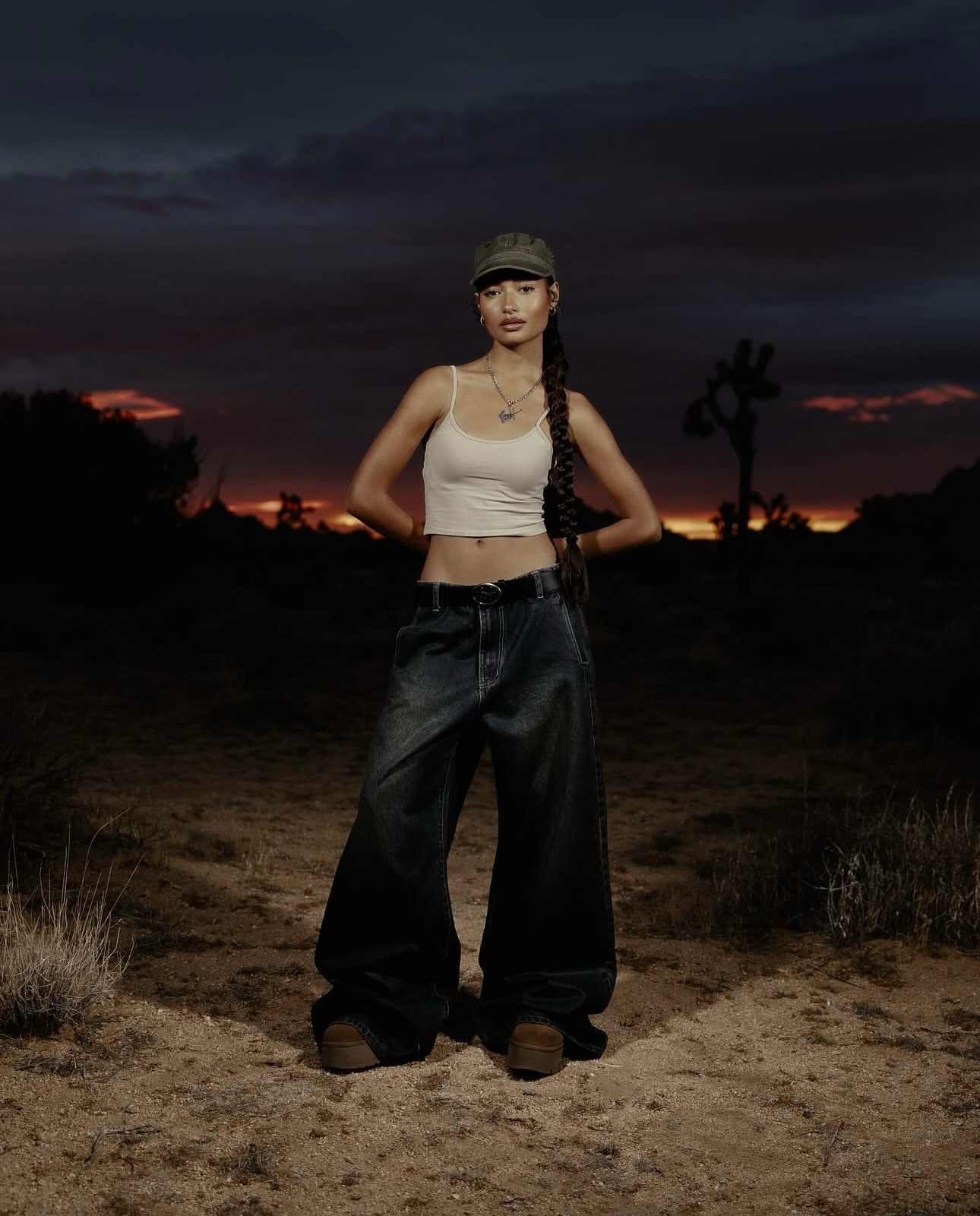 Young woman in casual streetwear stands in a desert landscape at dusk, wearing a tank top, baggy jeans, and a baseball cap.