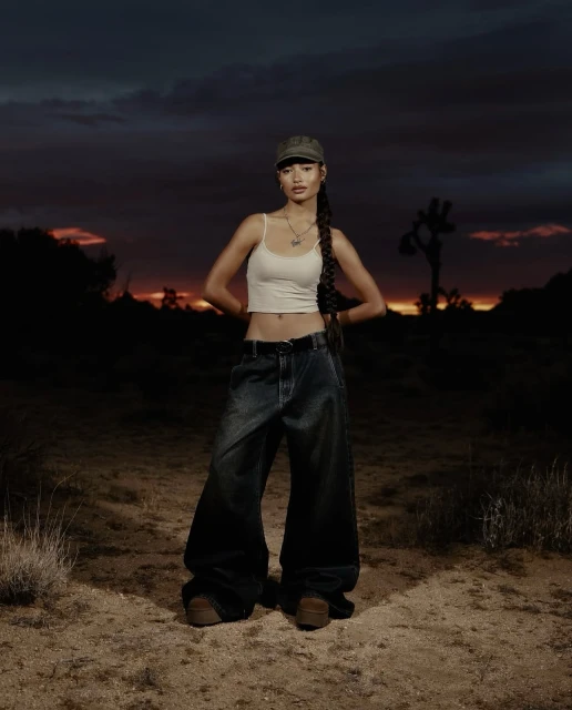 Young woman in casual streetwear stands in a desert landscape at dusk, wearing a tank top, baggy jeans, and a baseball cap.
