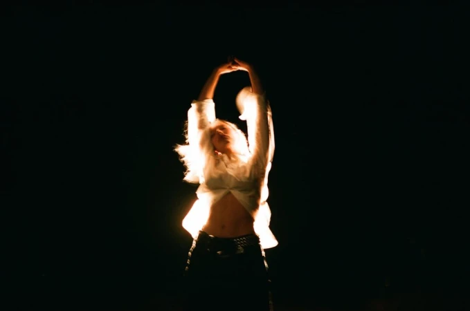 A woman in a white shirt with arms raised overhead, captured in dramatic low-key lighting against a dark background.