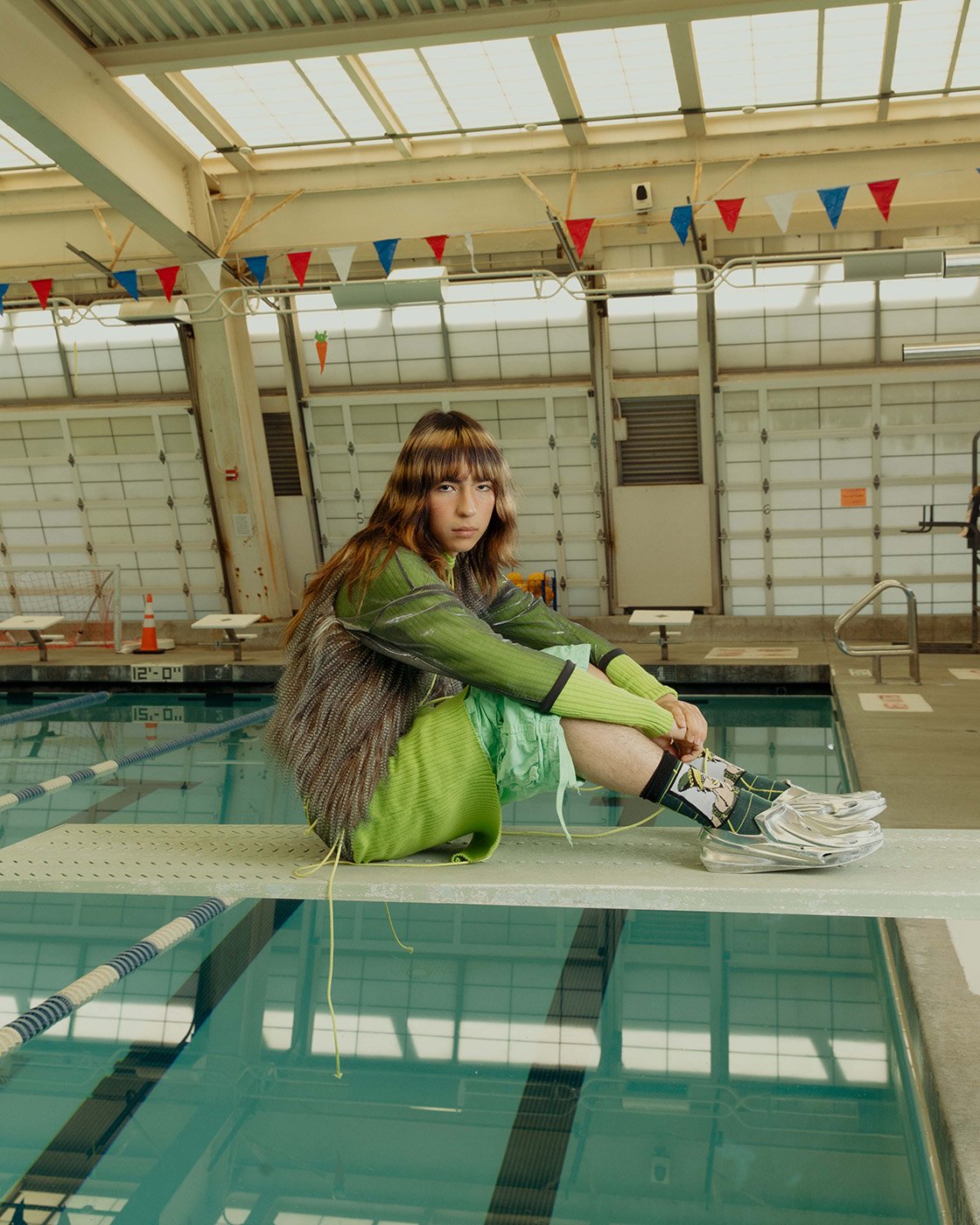 Young woman with bangs in a green outfit and fashion sneakers sitting on a diving board above a pool in an indoor swimming pool.