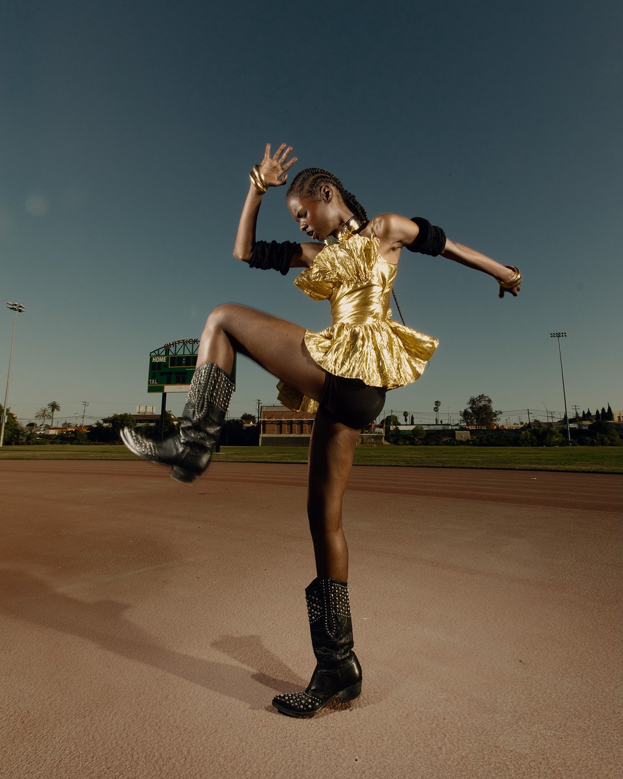 Athletic woman in gold top and studded boots strikes a pose on an outdoor track under a clear blue sky. Fashion editorial shot.