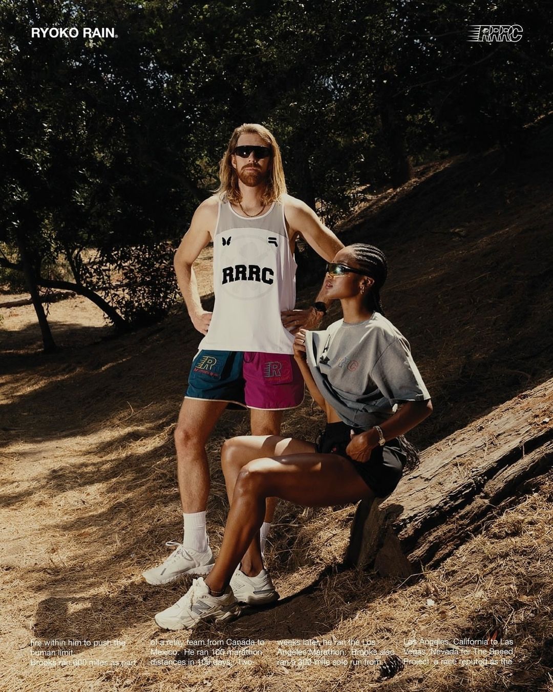 An athletic couple wearing sportswear stands on a trail in a sunlit forest. The woman sits on a fallen tree while the man stands.
