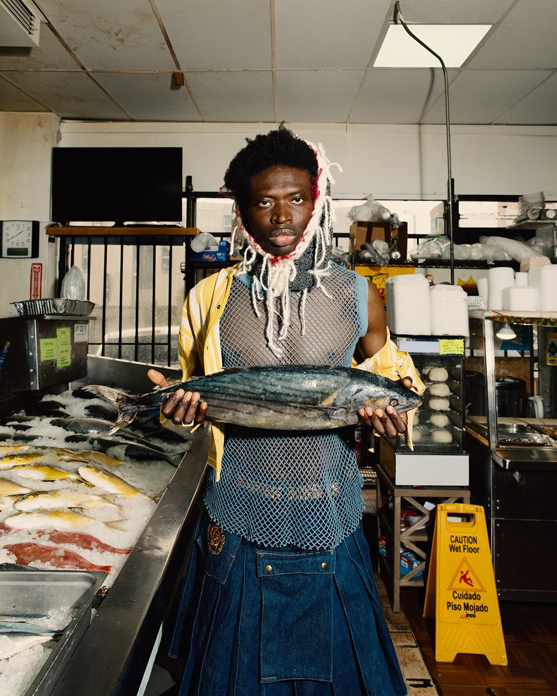 An adult holds a large fish in a fish market. The person wears a mesh top, denim skirt, and yellow jacket, standing among ice and other fish.