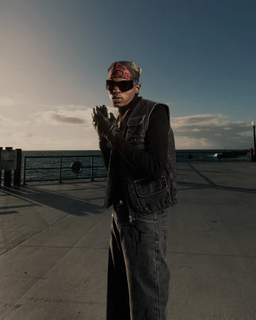 Man wearing a dark denim vest, black sunglasses, and a printed bandana standing outdoors near the ocean at sunset.