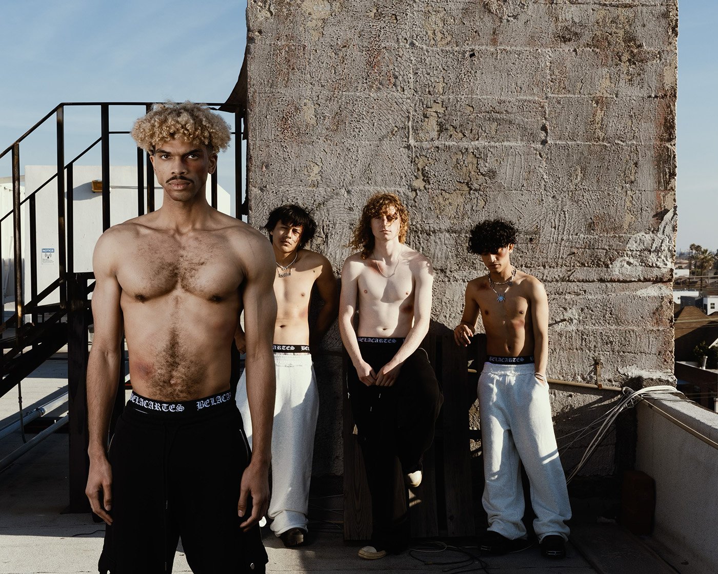 Group of four shirtless young adult men standing outdoors near a weathered concrete wall, wearing dark pants and light pants.
