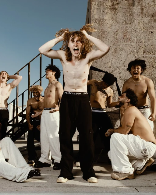 A group of men in casual streetwear poses outside with one man in the center pulling at his curly red hair and yelling.