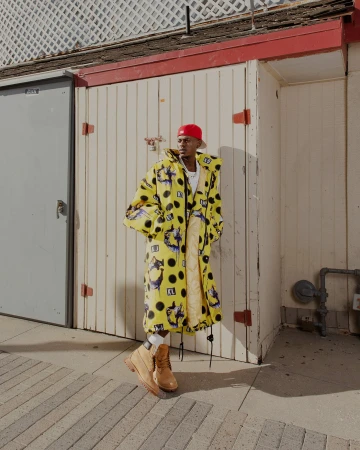 Black man wearing a yellow patterned puffer coat and red cap standing against a white wooden panel exterior wall.