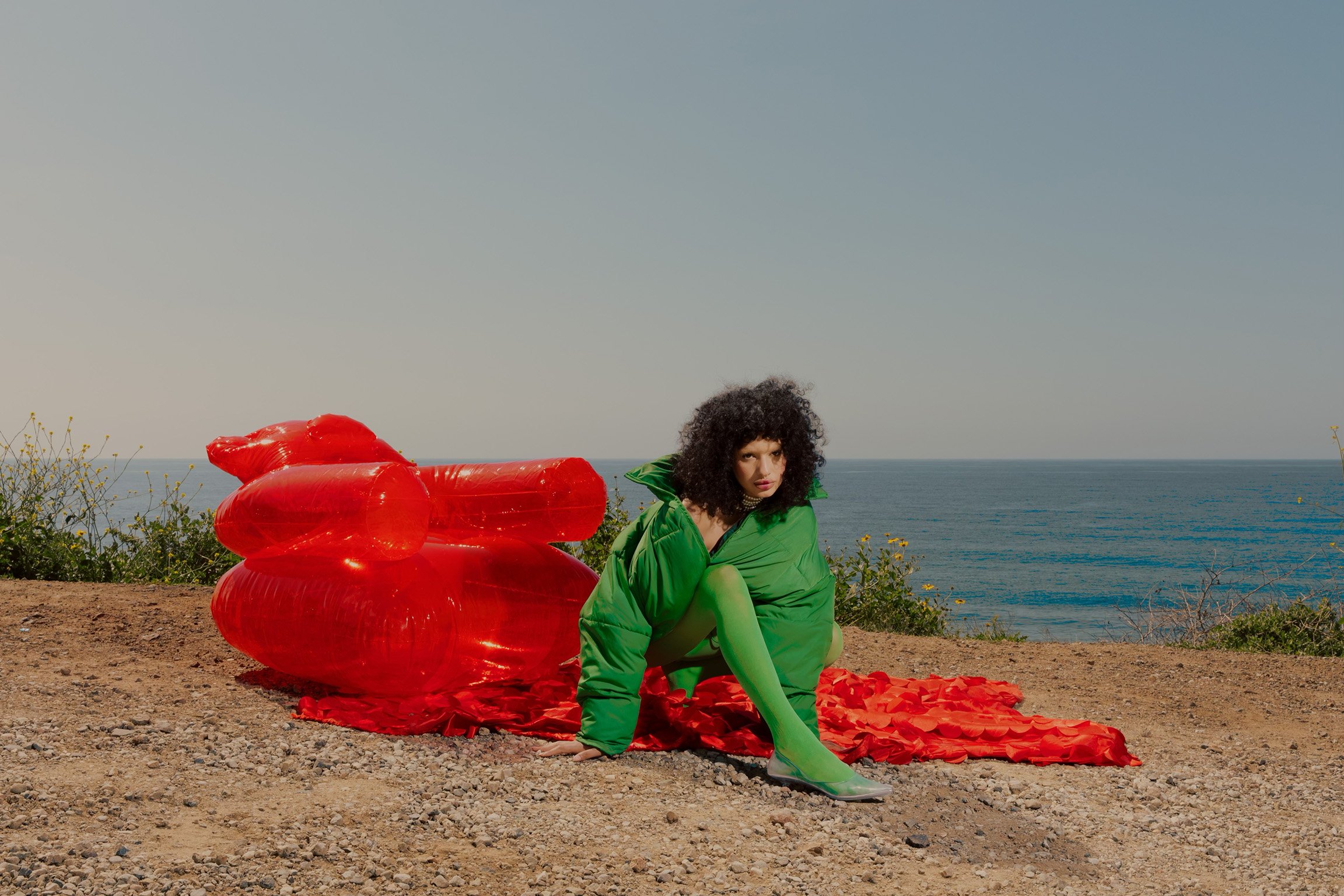 A woman in a green outfit sits near an inflatable red couch on a sandy bluff overlooking the ocean on a sunny day in a fashion editorial.