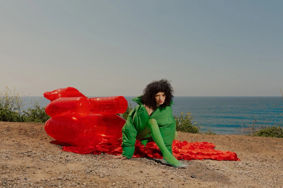 A woman in a green outfit sits near an inflatable red couch on a sandy bluff overlooking the ocean on a sunny day in a fashion editorial.