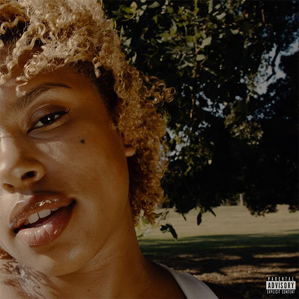 Close-up portrait of an adult woman with curly hair outdoors. She is viewed at eye-level, and a tree is visible in the background.
