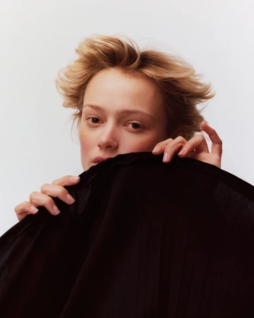 Close-up portrait of a woman with short blonde hair holding a dark fabric across the lower half of her face against a plain white background.