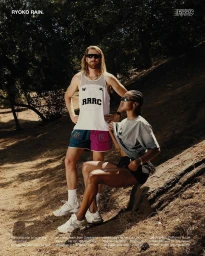 A man standing and a woman seated on a log in a wooded landscape, wearing athletic apparel and sunglasses.