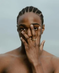 Portrait of a Black man with braided hair holding his hand in front of his face, wearing a gold ring.