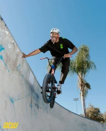 A Black man smiling while riding a BMX bike on a concrete skate ramp against a bright blue sky.