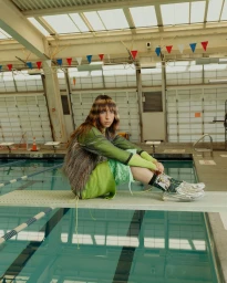 A person sitting on a diving board above an indoor swimming pool, wearing green and silver layered clothing.