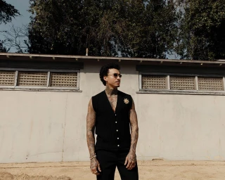 Man wearing a black sweater vest and dark sunglasses posing outdoors against a concrete building exterior in Los Angeles.